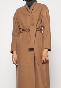 Persona che indossa un lungo cappotto avvolgente color cammello con una cintura annodata e un maglione dolcevita grigio scuro sotto.