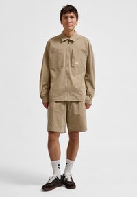 Beige ritsjack met twee voorzakken, gecombineerd met bijpassende shorts. Model draagt zwarte sneakers en witte sokken met zwarte strepen.