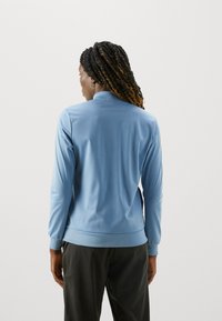 Svetlo modrá bunda na zips s vysokým golierom a dlhými rukávmi, vyrobená z hladkej tkaniny. Dolný lem a manžety majú rebrované akcenty.