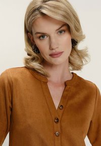 Hnědá semišová košile s kulatým límcem, třemi knoflíky vpředu a hladkým povrchem. Modelka má na sobě velké kruhové náušnice.