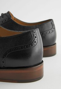 Chaussures en cuir noir avec une finition texturée, des détails brogue et un talon en bois. Présentent une silhouette élégante et des contours bien définis.