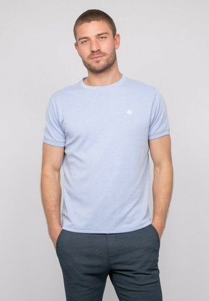 T-shirt bleu clair à manches courtes en tissu doux, avec un col rond et un petit logo sur la poitrine. Porté avec un pantalon texturé foncé.