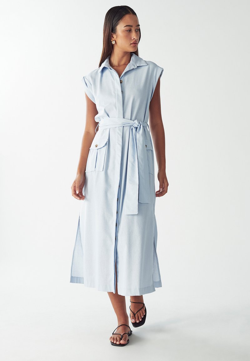 Vestito senza maniche di un blu cielo con chiusura frontale a bottoni, cintura in vita, due tasche a toppa e spacco laterale per il movimento.