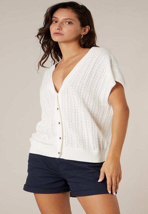 Femme aux cheveux ondulés foncés portant un cardigan blanc à manches courtes boutonné et un short bleu marine à ourlet retroussé, debout devant un fond uni.