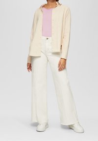 Beżowa koszula z guzikami nałożona na jasnoróżowy top, w połączeniu z szerokimi białymi dżinsami i białymi trampkami. Gładkie faktury, swobodny design.