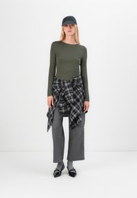 Olijfgroen langemouwen top, zwart-wit geruite blouse om de taille geknoopt, grijze wijde broek, grijze sokken, zwarte schoenen en een donkere pet.