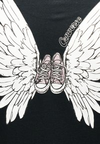 Ζεύγος ροζ αθλητικά παπούτσια Converse με λευκά φτερά που εκτείνονται από κάθε πλευρά, σε μαύρο φόντο με το κείμενο "Converse".