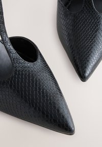 Fekete magas sarkú cipők, texturált, dombornyomott bőrből, hegyes orral és nyitott oldalsó kivágással.