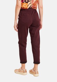 Donna con pantaloni viola scuri arrotolati, camicia a fantasia colorata e sneaker metallici rosa oro, in piedi con la schiena rivolta verso la macchina fotografica.