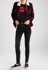 Chaqueta de franela a cuadros con cuello de borreguito blanco, camiseta negra con logo rojo, jeans ajustados negros y zapatillas negras sin cordones.