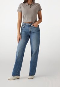 Kvinna iklädd en kort grå polo, högmidjade blå vida jeans och vita sneakers, står med en hand i fickan.