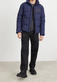 Marinblå puffjacka med dragkedja fram, logotyp på vänster bröst och hög krage. Svarta byxor och mörka skor fulländar outfiten.