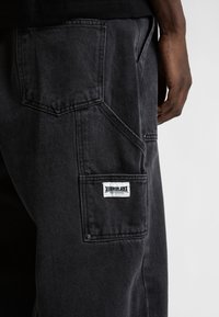 Juodi džinsiniai cargo kelnės su keliais kišenėmis, įskaitant didelę šoninę kišenę ir prekinį ženklą. Pasižymi tvirtu, tekstūruotu audiniu.