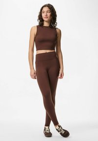 Femme en crop top sans manches marron et leggings assortis, les jambes croisées, portant des chaussures de sport marron et beige sur fond blanc.