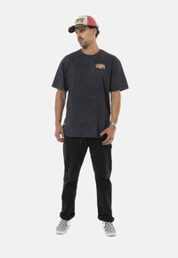 T-shirt surdimensionné gris foncé avec logo sur le devant, pantalon noir et chaussures à enfiler à carreaux. Une casquette rouge et beige complète la tenue.