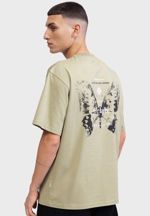 T-shirt in cotone verde oliva con una grande grafica di una farfalla nera sul retro e elementi testuali bianchi. Maniche corte e vestibilità comoda.