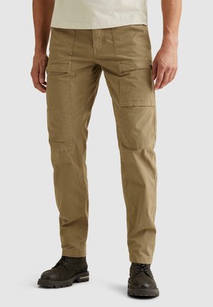 Mężczyzna ubrany w beżowe spodnie typu cargo z wieloma kieszeniami oraz czarne wiązane buty, stojący na tle jednolitego jasnego tła.