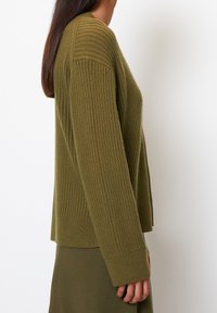 Žena v ribbeném olivově zeleném svetru a odpovídající sukni, stojící v profilu na pozadí z čistě bílé.