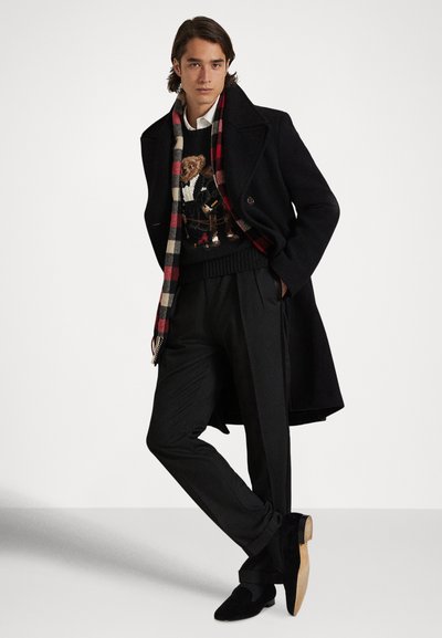 Άνδρας μοντέλο με μαύρο παλτό, μαύρο παντελόνι, μαύρο πουλόβερ με σχέδιο αρκουδάκι, άσπρο πουκάμισο, καρό κασκόλ και μαύρα παπούτσια, σε πόζα.