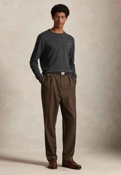 Giovane uomo in piedi con le mani in tasca, indossa una camicia grigio scuro a maniche lunghe, pantaloni a quadri marroni e scarpe in pelle marrone su sfondo neutro.