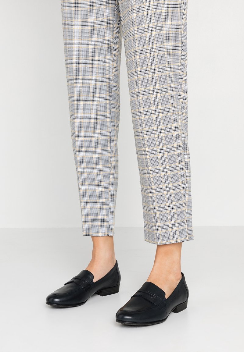Pantalon à carreaux bleu et beige longueur cheville, associé à des mocassins en cuir noir sur un fond clair.