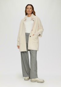 Lichtbeige oversized coat van textuurstof, met een kraag, twee voorkruimels en grote knopen, gecombineerd met grijze brede broekspijpen en beige laarzen.