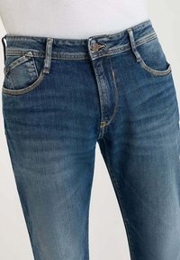 Jean en denim bleu foncé avec une coupe classique, comportant une poche avant, une fermeture à bouton en métal et des accents de couture visibles.