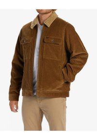 Mies, joka on pukeutunut ruskeaan corduroy-takkiin, jossa on sherpa-kaulus, valkoiseen paitaan ja beigeihin housuihin, käsi takin taskussa, seisoo valkoista taustaa vasten.