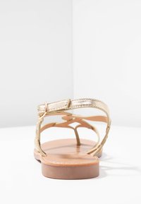 Sandali metallici dorati con design a strisce incrociate e una suola piatta color tan. Presentano una chiusura a fibbia e dettagli testurizzati sulle cinghie.