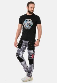 Schwarzes Baumwoll-T-Shirt mit grafischem Design auf der Vorderseite. Graue Cargo-Hosen mit Marmor-Muster, mehreren Taschen und schwarzen Akzenten. Weiße Sneakers.