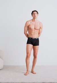 Schwarze Boxershorts mit elastischem Bund, der das "HUGO"-Logo trägt. Das Modell hat eine muskulöse Statur und steht barfuß auf einer hellen, strukturierten Fläche.