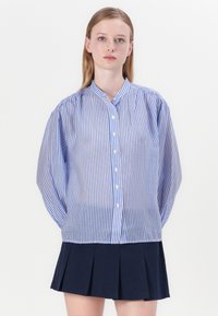 Sininen ja valkoinen raidallinen napillinen paita, jossa on tuki kaulus ja puffihihat, yhdistettynä tummansiniseen laskostettuun hameeseen.