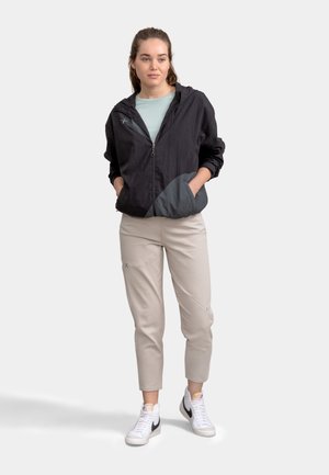 Zwarte lichtgewicht jas met grijze accenten, ritssluiting aan de voorkant en zijzakken, gecombineerd met lichtbeige broek en witte sneakers.