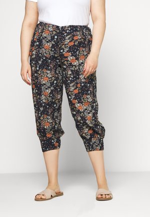 Kobieta w czarnych skróconych spodniach w pomarańczowo-biały kwiatowy wzór, białej koszuli i beżowych sandałach wsuwanych, stojąca na białym tle.