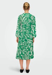 Abito midi verde con motivo floreale bianco, maniche lunghe e design a strati. Modella che indossa scarpe basse nere.