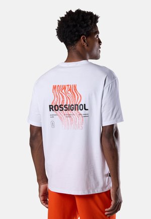 Mężczyzna w białej koszulce Rossignol z czerwono-czarnym nadrukiem na plecach o tematyce górskiej, w połączeniu z jasnopomarańczowymi szortami.