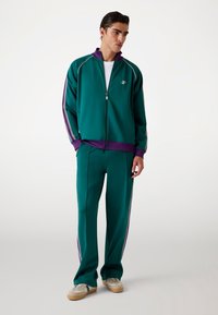 Teal väzipälsjoggingdress med lila detaljer, som har kontrasterande sidostreck och avslappnade byxor. Tillverkad av ett mjukt, stretchigt material.