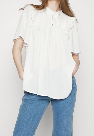 Vrouw die een losse witte blouse met plooien en ruches aan de mouwen draagt, gecombineerd met een blauwe spijkerbroek, handen in de zakken, staand tegen een effen achtergrond.