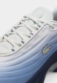 Λευκό αθλητικό παπούτσι με άνω μέρος από δίχτυ, λευκά κορδόνια, γκρι θηλιές για τα κορδόνια και μικρό λογότυπο κροκόδειλου στο πλάι κοντά στη σόλα.