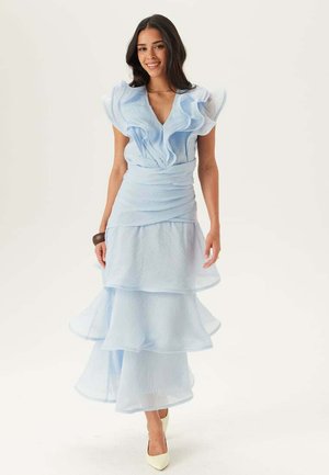 Donna sorridente che indossa un vestito azzurro chiaro a balze con scollo a V profondo e tacchi bianchi appuntiti, in piedi davanti a uno sfondo bianco uniforme.