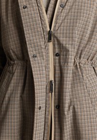 Close-up van een bruin- en beige geruit jack met houndstooth-patroon, zwarte drukknoopsluitingen en een beige rits, met zijzakken en een elastische taille.