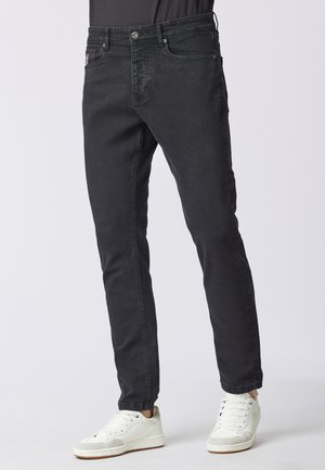 Jeans in denim nero con vestibilità slim, design a cinque tasche, cuciture sottili e chiusura con bottone, abbinati a sneakers bianche.