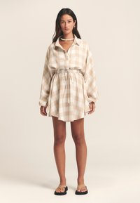 Hellbeiges kariertes Kleid mit lockerem Schnitt, langen Ärmeln und verstellbarem Tunnelzug in der Taille. Kombiniert mit schwarzen Flip-Flop-Sandalen.