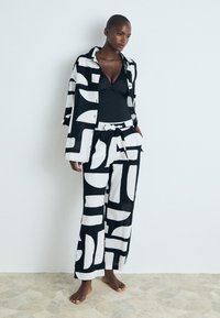 Črno-belo vzorčasta lanena srajca in široke hlače, ki vključujejo abstraktne oblike, pas s povezovanjem in plitko ovratnikom.