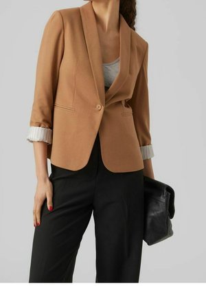 Kobieta w camelowym blezerze z podwiniętymi, paskowanymi mankietami, nosząca szarą bluzkę, czarne spodnie, trzymająca czarną skórzaną kopertówkę.