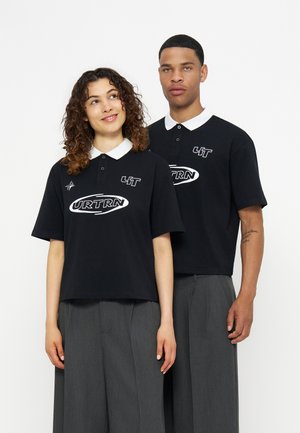 Deux personnes portant des polos noirs avec des cols blancs et des pantalons gris à jambes larges, debout devant un fond blanc uni.