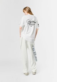 Camiseta blanca oversize con letras negras en la parte posterior, combinada con pantalones blancos holgados que tienen una etiqueta gráfica en el costado.