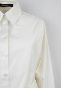 Camicia bianca con colletto classico, caratterizzata da bottoni trasparenti e un tessuto testurizzato. Include una tasca sulla parte anteriore.