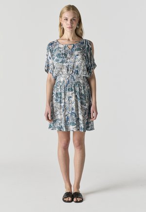 Mujer vistiendo un vestido de manga corta con un estampado azul y blanco, con aberturas en los hombros y sandalias negras sin cierre, de pie sobre un fondo liso.