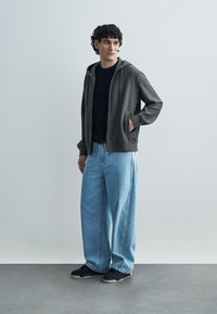 Γκρι φούτερ με φερμουάρ, με μπροστινές τσέπες, συνδυασμένο με άνετο, φαρδύ τζιν σε ανοιχτό γαλάζιο χρώμα. Μαύρα αθλητικά παπούτσια ολοκληρώνουν το σύνολο. Ομαλή υφή, καθημερινός σχεδιασμός.